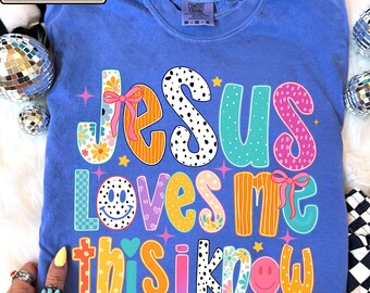 May include: A periwinkle blue t-shirt featuring the colorful text JESUS LOVES ME THIS I KNOW in various patterns including floral, polka dots, stripes, and checkerboard. The design includes pink bows and smiley faces on a cotton fabric.