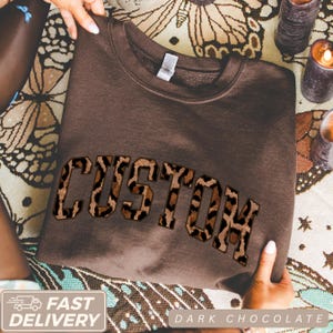 Könnte beinhalten: Ein dunkel schokoladenbraunes Sweatshirt mit Rundhalsausschnitt und dem Wort CUSTOM in großen Blockbuchstaben auf der Brust. Die Buchstaben haben ein braun-schwarzes Leopardenmuster mit dunkler Umrandung, was ein auffälliges Design erzeugt.