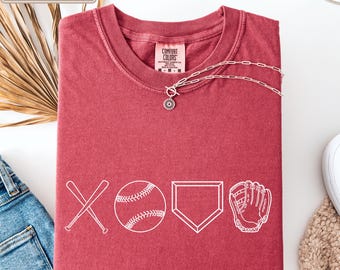 May include: Crimson-colored t-shirt with white baseball-themed graphics. The design includes crossed bats, a baseball, home plate, and a baseball glove. The shirt has a crew neck and short sleeves. A silver necklace with a pendant is on the shirt.