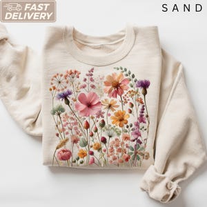 May include: A sand-colored crewneck sweatshirt featuring a central graphic print of various wildflowers in shades of pink, orange, purple, and green. The design includes detailed illustrations of cosmos, cornflowers, and assorted meadow grasses.