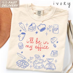 以下が含まれることがあります： アイボリー色のTシャツ。牛乳パック、シェフハット、小麦粉袋、オーブンミトン、カップケーキ、麺棒、卵、エプロン、秤などの製菓道具の青い線画イラストが描かれており、中央には赤い文字で「i'll be in my office」と記されています。