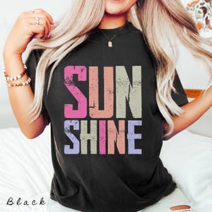 May include: Black t-shirt with the word "SUNSHINE" in a distressed, multi-colored font. The word "SUN" is in pink, peach, and beige, while "SHINE" is in purple, pink, and peach. The word "Black" is written in cursive at the bottom.