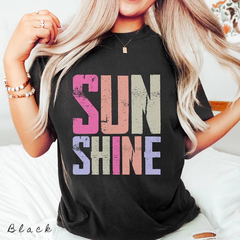 May include: Black t-shirt with the word "SUNSHINE" in a distressed, multi-colored font. The word "SUN" is in pink, peach, and beige, while "SHINE" is in purple, pink, and peach. The word "Black" is written in cursive at the bottom.