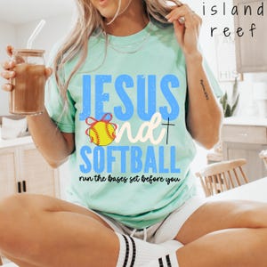 May include: A mint green short-sleeve t-shirt featuring the blue text JESUS and SOFTBALL with a yellow softball graphic tied with a red ribbon. The shirt includes the black text run the bases set before you and a small black cross symbol.