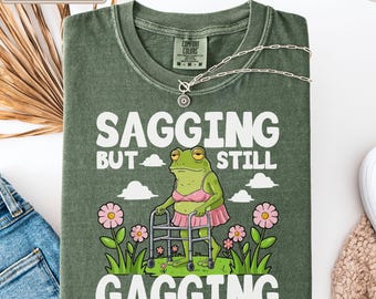 Op de afbeelding: Een mosgroen t-shirt met een afbeelding van een kikker in een roze bh en rok die een rollator gebruikt. De tekst luidt "SAGGING BUT STILL GAGGING" met illustraties van roze bloemen en witte wolken rondom de kikker op een groen vlak.