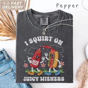 Op de afbeelding: Een antracietgrijs t-shirt met een afbeelding van een cartoon ketchupfles en een hotdog. De tekst luidt I SQUIRT ON JUICY WIENERS en LOVE JUICE op de fles. Het ontwerp bevat bloemen, wolken en sterren in een retrostijl op de voorzijde.