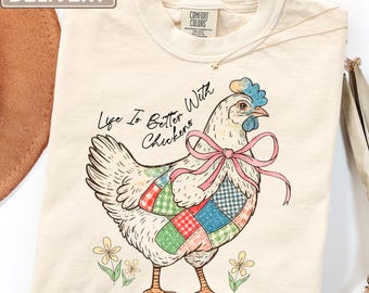 Może przedstawiać: T-shirt w kolorze kości słoniowej z grafiką białej kury w różowej kokardzie i kolorowej kamizelce patchworkowej. Nad kurą znajduje się napis "Life Is Better With Chickens", a na dole umieszczono ilustracje małych żółtych kwiatków.