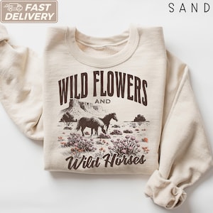May include: A sand-colored crewneck sweatshirt featuring a dark brown graphic print. The design displays a desert landscape with mountains, two horses, and wildflowers, accompanied by the text "WILD FLOWERS AND Wild Horses" in a bold, arched font.
