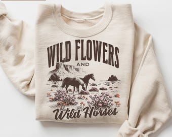 May include: A sand-colored crewneck sweatshirt featuring a dark brown graphic print. The design displays a desert landscape with mountains, two horses, and wildflowers, accompanied by the text "WILD FLOWERS AND Wild Horses" in a bold, arched font.