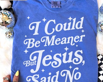 Pode incluir: Uma t-shirt azul de manga curta com texto serigrafado em branco que diz I Could Be Meaner But Jesus, Said No, acentuado com pequenos gráficos de estrelas brancas. A t-shirt apresenta um decote redondo e uma etiqueta de tamanho medium.