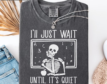 May include: A charcoal gray short-sleeve t-shirt featuring a white graphic of a skeleton sitting at a chalkboard with the text I'LL JUST WAIT UNTIL IT'S QUIET. The shirt has a crew neckline and a Comfort Colors brand label printed on the inner collar.