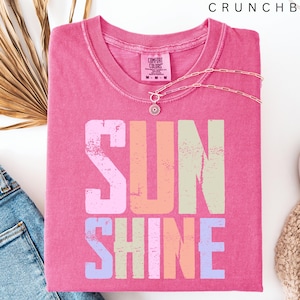 May include: A pink t-shirt with the word "SUNSHINE" in a colorful, distressed font. A delicate silver necklace with a pendant rests on the shirt. The shirt is paired with blue jeans, a white shoe, and a beige knit item.