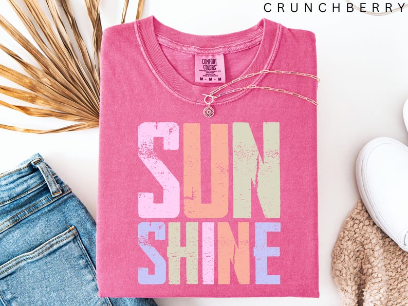 May include: A pink t-shirt with the word "SUNSHINE" in a colorful, distressed font. A delicate silver necklace with a pendant rests on the shirt. The shirt is paired with blue jeans, a white shoe, and a beige knit item.