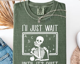 Può includere: Una t-shirt a girocollo color verde muschio con una grafica bianca raffigurante uno scheletro seduto davanti a una lavagna con il testo I'LL JUST WAIT UNTIL IT'S QUIET. La maglia presenta un'etichetta Comfort Colors sul colletto e una catenina.