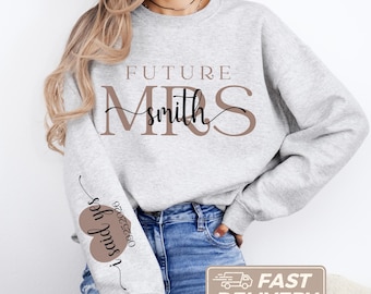 Könnte beinhalten: Ein hellgraues Sweatshirt mit Rundhalsausschnitt und dem Text "FUTURE MRS" in großen braunen Buchstaben sowie dem Wort "smith" in schwarzer Schreibschrift in der Mitte. Der Ärmel zeigt ein braunes Herz mit "i said yes" und "09.25.2026".