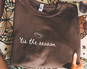 May include: A dark brown crewneck sweatshirt featuring white embroidered text that reads 'tis the season with a small white embroidered football icon positioned directly above the text on the centre chest area.
