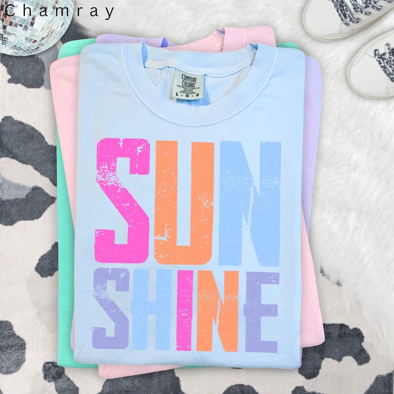 May include: A stack of pastel-colored t-shirts. The top shirt is light blue with the word "SUNSHINE" printed in distressed pink, orange, and blue. The shirts are folded neatly, with a tag visible on the top shirt. The shirts are on a white and black cow print surface.