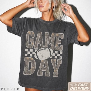 Pode incluir: Uma t-shirt de manga curta cinzento-carvão com o texto GAME DAY em letras grandes com um padrão de camuflagem. Um gráfico de uma bola de futebol americano está centrado entre as palavras, acompanhado por um design de borda xadrez.