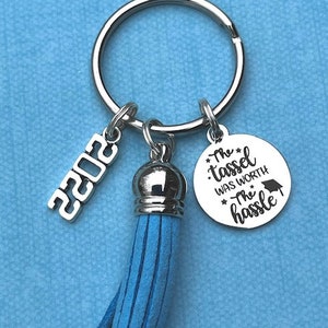 May include: A silver keychain with a blue tassel, a silver charm with the text "The tassel was worth the hassle" and a silver charm with the year "2022".