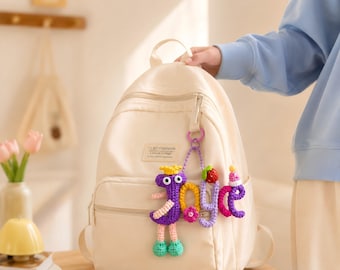 Könnte beinhalten: Cremefarbener Rucksack mit einem gehäkelten Schlüsselanhänger. Der Schlüsselanhänger zeigt eine lila vogelförmige Figur, das Wort "joyce" in bunten Buchstaben und eine Erdbeere. Der Rucksack hat einen Reißverschluss und ein Etikett "CLRD FASHION".