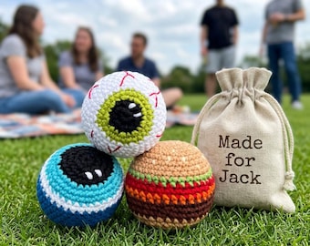 Könnte beinhalten: Drei gehäkelte Bälle: ein Augapfel, ein blau-weiß gestreifter Ball und ein Burger. Ein Kordelzugbeutel mit "Made for Jack" befindet sich neben den Bällen. Die Bälle liegen auf grünem Gras mit Personen im Hintergrund.