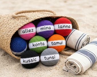 Könnte beinhalten: Ein geflochtener Strohbeutel gefüllt mit bunten, gehäkelten Bällen mit weißen Streifen und verschiedenen Namen. Ein aufgerolltes gestreiftes Handtuch liegt am Sand neben der Tasche. Die Bälle sind in den Farbtönen Rosa, Lila, Rot, Blau, Grün, Orange, Schwarz und Grau.