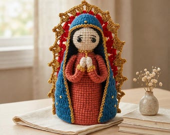 Könnte beinhalten: Handgefertigte Häkelpuppe, die die Jungfrau Maria darstellt. Die Puppe hat ein rotes Kleid, einen blauen Umhang mit goldenen Verzierungen und Sternen, sowie einen goldenen Heiligenschein. Die Puppe steht auf einem weißen Tuch, mit einer kleinen Vase mit Blumen im Hintergrund.
