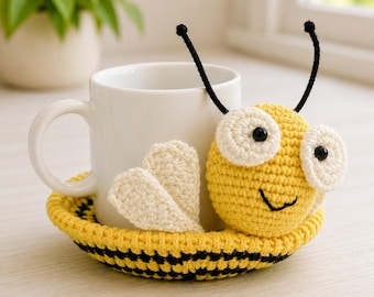 Könnte beinhalten: Eine weiße Tasse steht auf einem handgefertigten Bienen-Untersetzer aus Häkelarbeit. Der Untersetzer hat einen gelben Körper mit schwarzen Streifen, weiße Flügel und ein lächelndes Gesicht. Schwarze Antennen vervollständigen das Design.