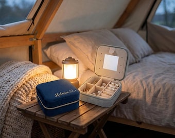Könnte beinhalten: Zwei Schmuckkästchen stehen auf einem Holztisch. Eines ist marineblau mit einem goldenen Reißverschluss und dem Namen "Hilaria" in Gold gestickt. Das andere ist offen, hellgrau und enthält Schmuck. Eine kleine Laterne und ein Bett sind im Hintergrund.
