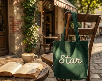 Könnte beinhalten: Eine grüne Cord-Tragetasche mit dem Wort "Star" in Weiß gestickt. Die Tasche hängt an einem Holzstuhl. Eine Tasse Kaffee und ein offenes Buch liegen auf einem Holztisch vor einem Café.
