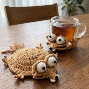 May include: Two brown crocheted squirrel-shaped coasters with large white eyes and black pupils. One coaster holds a clear glass mug filled with tea. The coasters are on a wooden table.