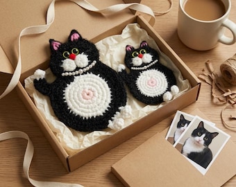 Könnte beinhalten: Ein Set aus schwarz-weißen Häkel-Katzenuntersetzern mit weißem Bauch und rosa Akzenten, präsentiert in einer Pappschachtel. Das Set enthält zwei Untersetzer und zwei Katzenfotos. Eine Tasse Kaffee steht daneben.