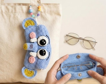 Könnte beinhalten: Ein hellblaues, gehäkeltes Brillenetui in Form eines Cartoon-Monsters mit großen schwarzen und weißen Augen, rosa Akzenten und gelben Details. Eine runde Brille und ein passendes Etui sind ebenfalls zu sehen.