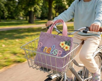 Könnte beinhalten: Eine lavendelfarbene Cord-Tragetasche mit dem Namen "Penelope" in bunten Buchstaben gestickt und zwei gelben Blumenapplikationen. Die Tasche hat rosa Griffe und befindet sich in einem Fahrradkorb. Das Fahrrad wird auf einem Weg gefahren.