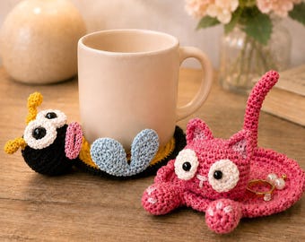 Könnte beinhalten: Eine beige Keramiktasse steht auf einem handgefertigten Häkeluntersetzer mit einem schwarzen, gelben und blauen Design. Ein rosa Katzen-Häkel-Tablett mit einem Ring und Perlen befindet sich in der Nähe.