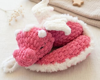 Könnte beinhalten: Ein handgefertigtes, gehäkeltes Drachenspielzeug in Rosa und Weiß. Der Drache hat weiße Flügel, einen weißen Bauch und weiße Akzente an Kopf und Schwanz. Der Drache liegt auf einer weißen Oberfläche.