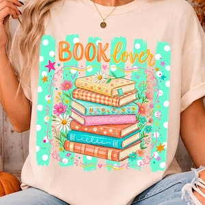May include: A cream-colored short-sleeve t-shirt featuring a graphic print of a stack of six colorful books. The text "BOOK lover" is printed in orange above the books, which are surrounded by illustrations of flowers, stars, and dots.