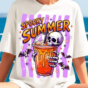 May include: A white t-shirt featuring a graphic print of a skeleton hand holding an orange iced beverage with a skull resting on top. The text "SPOOKY SUMMER" is printed above the drink in orange and yellow, surrounded by purple stripes and bats.