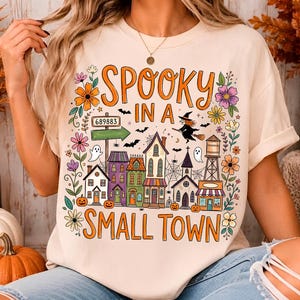 May include: A cream-colored t-shirt featuring a colorful graphic with the text "SPOOKY IN A SMALL TOWN". The design includes a row of houses, a water tower, a witch on a broomstick, ghosts, bats, spiderwebs, and various orange and purple flowers.