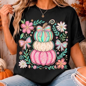 Pode incluir: Uma t-shirt preta de manga curta com uma estampa gráfica de três abóboras empilhadas em tons de turquesa, branco e rosa com padrões florais. O design inclui laços em tons pastel, margaridas e ramos verdes à volta das abóboras.