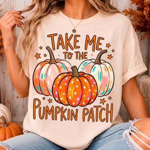 Peut inclure: Un t-shirt couleur crème avec un motif de trois citrouilles aux accents orange, rose et bleu. Le texte "TAKE ME TO THE PUMPKIN PATCH" est écrit en lettres orange, entouré de petites étoiles orange. Le motif est centré sur le devant du t-shirt.