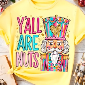 May include: A light yellow t-shirt featuring a colorful nutcracker graphic with a gold glitter heart on its hat. The text reads Y'ALL ARE NUTS in pink and blue lettering. The design includes small gold stars and snowflakes on a yellow background.
