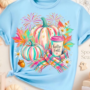 May include: A light blue t-shirt featuring a colorful graphic of two pumpkins, a plaid scarf, and a coffee cup with the text "hello Fall" and a small heart. The design includes autumn leaves, daisies, and fireworks in vibrant pink, orange, and teal.