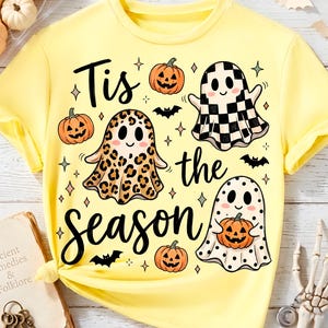 May include: A pale yellow t-shirt featuring a graphic print with the text "Tis the season" in black script. The design includes three ghosts with different patterns, including leopard print, checkerboard, and polka dots, alongside orange pumpkins and bats.