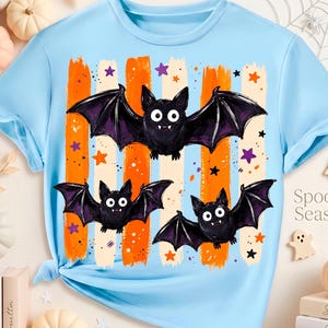 May include: A light blue t-shirt featuring a graphic of three black bats with white eyes and fangs. The design includes vertical orange and cream brushstroke stripes, small purple and orange stars, and the text Spooky Season on the side.