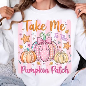 Può includere: Una felpa bianca a girocollo con una stampa grafica che riporta il testo "Take Me To The Pumpkin Patch". Il design presenta tre zucche nei toni dell'arancione, del rosa e del giallo, decorate con fiocchi, stelle e foglie su uno sfondo a righe rosa.