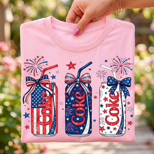 Pode incluir: Uma t-shirt rosa claro com um gráfico de três latas de refrigerante com palhinhas e laços. As latas são decoradas com padrões de bandeira vermelha, branca e azul, estrelas e fogo de artifício. A palavra Coke está impressa a vermelho em cada lata.