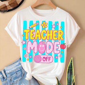 May include: White t-shirt with a colorful graphic design. The design features the words "TEACHER MODE" in yellow and pink, along with a smiley face, an apple, a pencil, and stars. The shirt has blue vertical stripes.