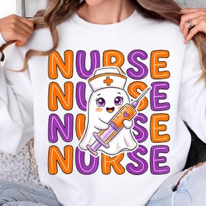 May include: A white sweatshirt featuring a repeating graphic of the word NURSE in orange and purple block letters. In the center, a cartoon ghost wearing a nurse cap holds a large syringe filled with orange liquid and a small purple heart.