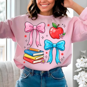 May include: A light pink sweatshirt featuring colorful school-themed graphics. The design includes a pink bow, a red apple, a blue bow, and a stack of books with a pencil. The graphics are surrounded by stars and small diamonds.